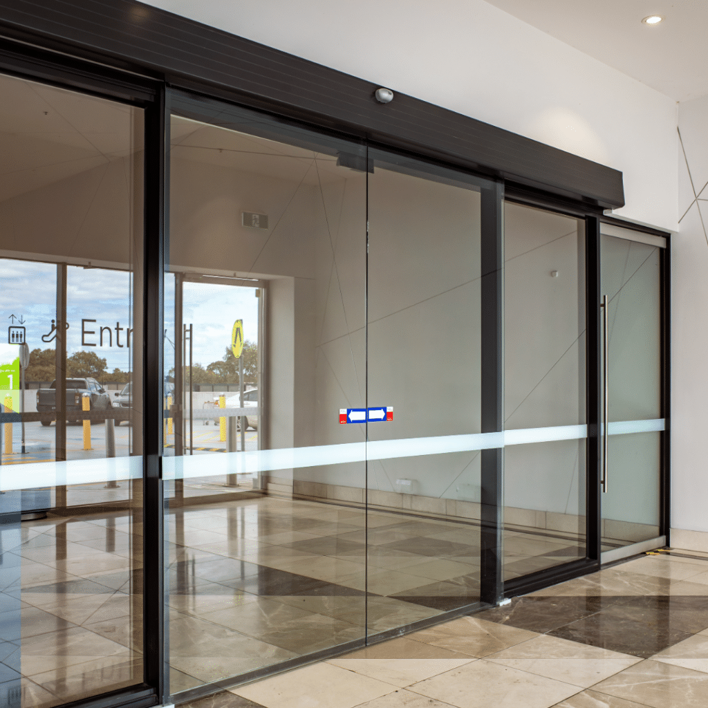 Image of automatic sliding doors on a building, black frames with white wall surrounding it. Why automatic door maintenance matters blog by J Manny.